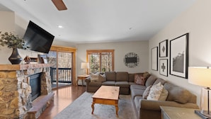 A spacious living room with a stone fireplace, a large sectional sofa, a wooden coffee table, and a flat-screen TV mounted on the wall. - Short Walk to Gondola, Shops, Dining, Mountain Views from Private Balcony! (Keystone)