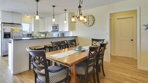 Een keuken met een centraal eiland, een houten eettafel en stoelen. Er hangt een klok aan de muur en bevindt zich een deur aan de rechterkant.