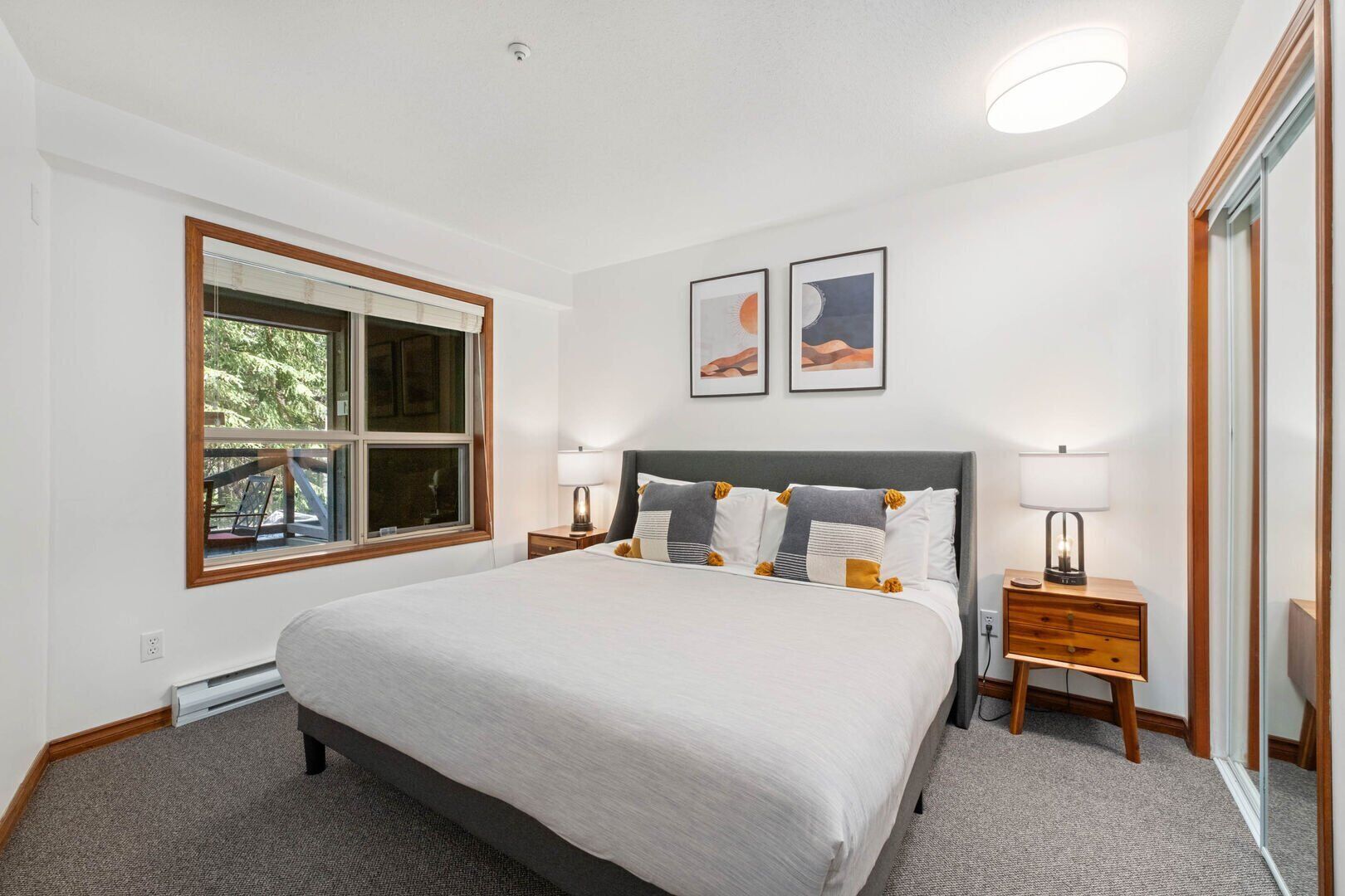 A bedroom with a large bed, two bedside tables with lamps, a window with blinds, and two framed pictures on the wall.