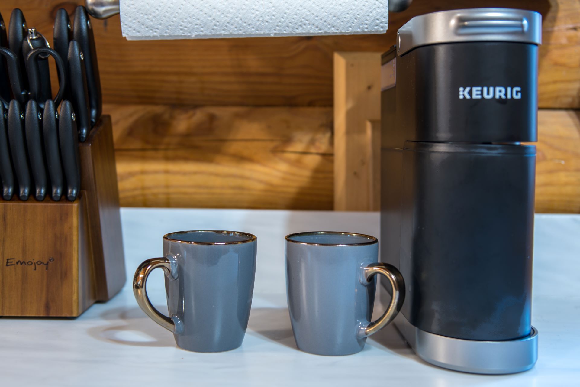 A Keurig coffee maker, two grey mugs, and a knife block on a wooden surface.