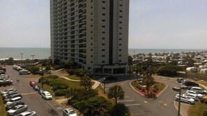 A high-rise building with a parking lot in front, surrounded by greenery and a view of the ocean.