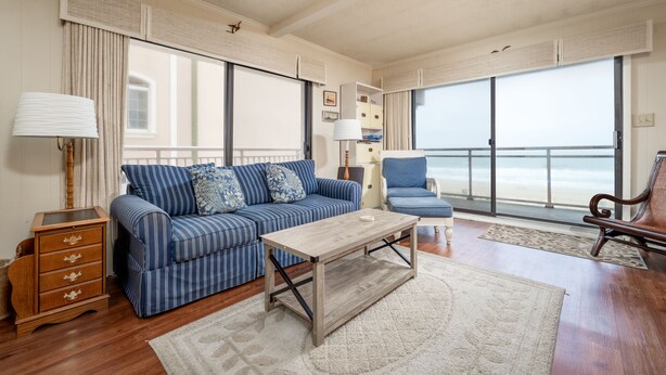 A living room with a striped sofa, a wooden coffee table, a blue armchair, and a view of the ocean through large windows. - Two bedroom oceanfront condo, just two blocks to the boardwalk! (Ocean City)