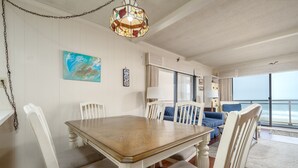 A dining area with a wooden table and white chairs, a large window with a view, and a hanging stained glass light fixture. - Two bedroom oceanfront condo, just two blocks to the boardwalk! (Ocean City)