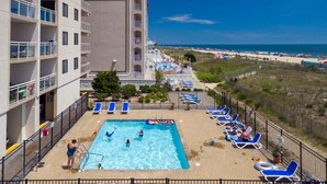 A pool area with a view of the beach, lounge chairs, and a building with multiple balconies. - Two bedroom oceanfront condo, just two blocks to the boardwalk! (Ocean City)