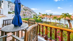Un balcon avec une table et des chaises en bois, un parasol bleu et une vue sur une colline parsemée de bâtiments et de palmiers.