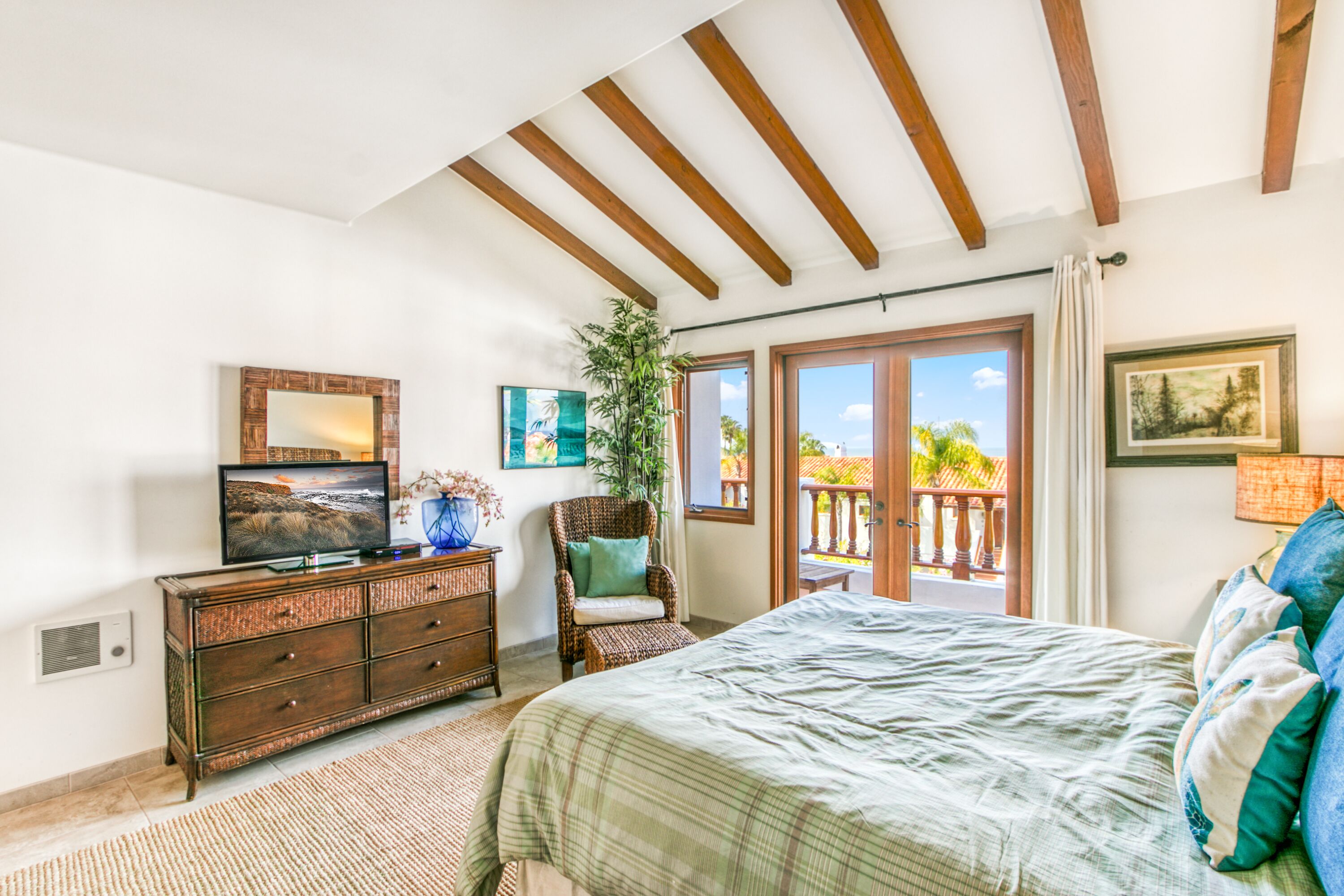 Ein Schlafzimmer mit einem Bett, einem Fernseher auf einer Holzkommode, einem Korbstuhl und einem Balkon mit Blick auf Palmen.