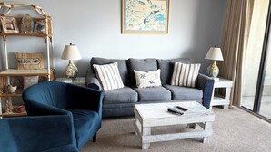 Una sala de estar con un sillón azul, sofá gris y una mesa de centro. Hay dos lámparas en mesas auxiliares, un cuadro en la pared y un estante de mimbre con objetos decorativos.