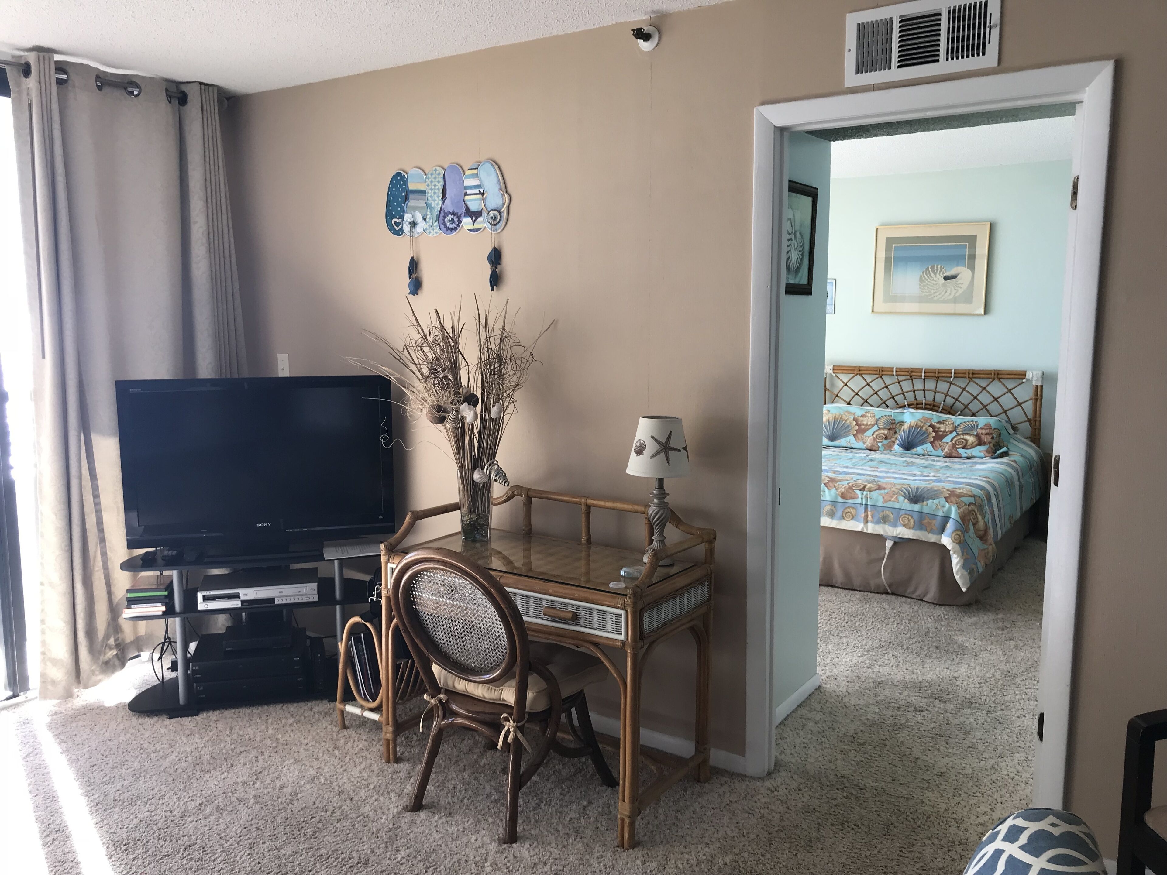 A room with a television, a desk with a lamp, a chair, and a bed with a blue and white patterned bedspread.
