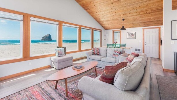 Ein geräumiges Wohnzimmer mit großen Fenstern, die auf einen Strand blicken, einer Holzbalkendecke und einer gemütlichen Sitzecke.
