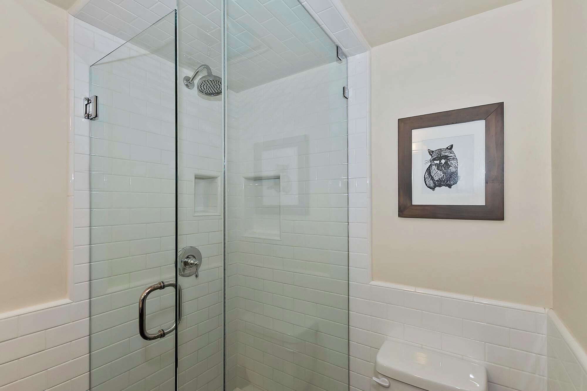 A glass-enclosed shower with a handheld showerhead, a framed picture of a cat on the wall, and a white tiled sink.