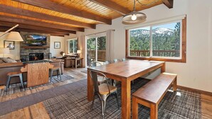 L’intérieur chaleureux d’un chalet avec une table à manger en bois, des bancs, une cheminée, un téléviseur et une grande fenêtre donnant sur une vue imprenable sur les montagnes.