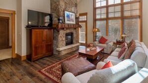 A living room with a stone fireplace, a television, and a large window with blinds. - Ski-In/Ski-Out Penthouse Condo w/ Large Balcony for Gorgeous Slope Views! (Keystone)