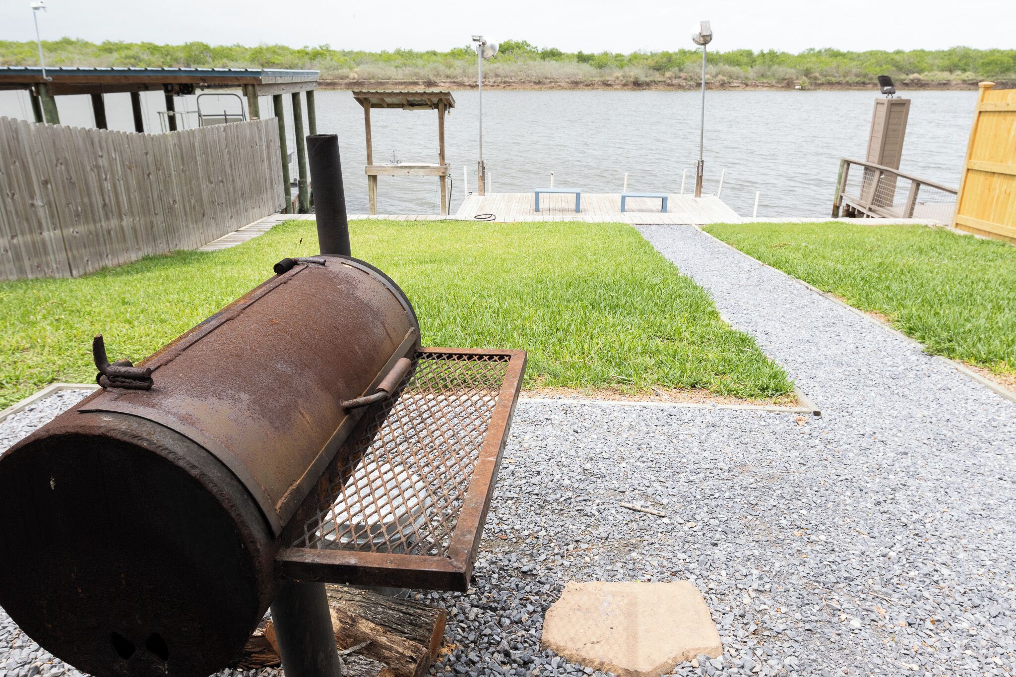 A barbecue grill outdoors with a wooden fence, a dock, and a body of water in the background.