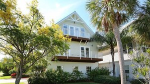 A two-story house with a balcony, surrounded by lush greenery and palm trees.