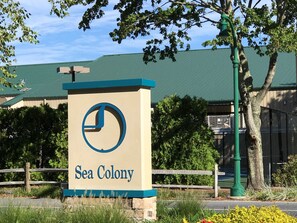 A sign for Sea Colony with a clock symbol, surrounded by greenery and a fence. - Sea Colony, Walk to Beach & Tennis--Beautiful, Sparkling Clean Getaway! (Bethany Beach)