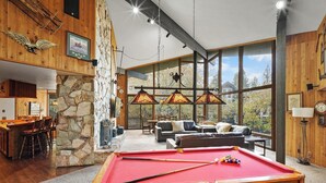 Un amplio salón con mesa de billar, chimenea de piedra y grandes ventanales con vistas al exterior.