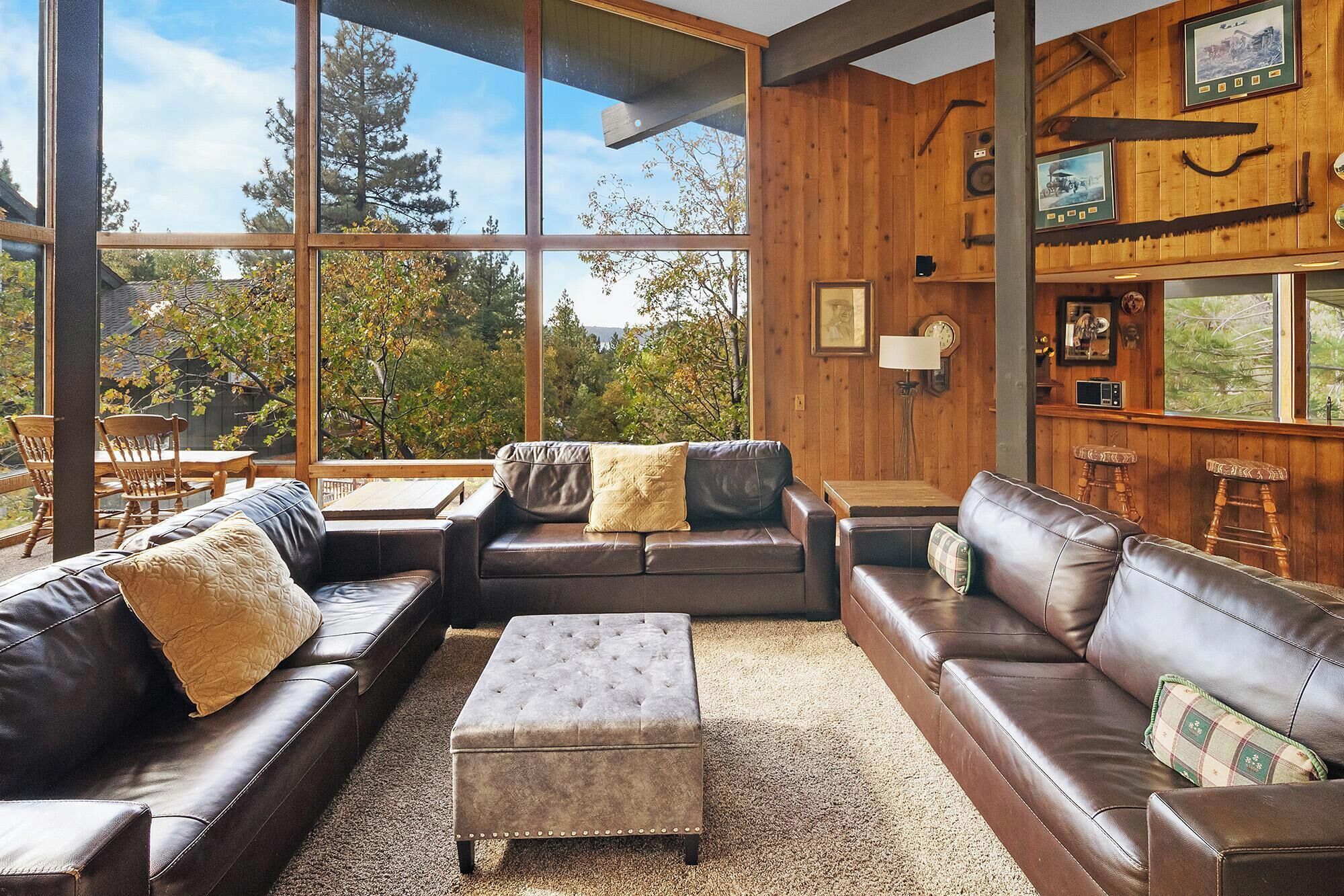 A spacious living room with leather sofas, a tufted ottoman, and large windows offering a view of trees and sky.