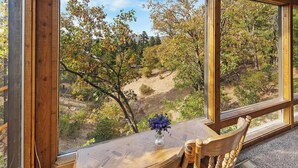 Una mesa de madera con un florero, frente a una vista panorámica de árboles y colinas a través de amplios ventanales.
