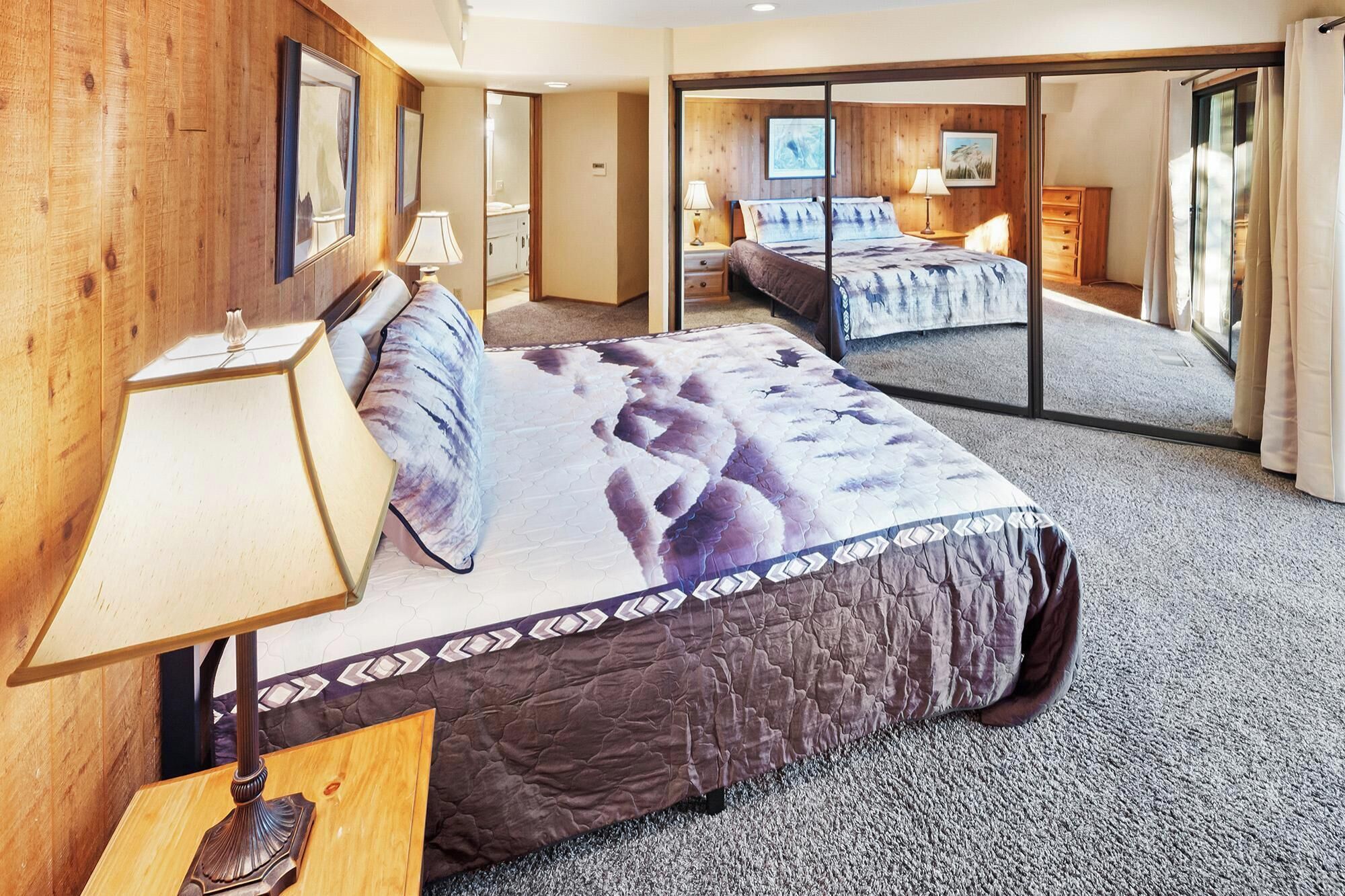 Habitación de hotel con una cama grande, cabecera de madera, mesita de noche, lámpara y espejo.