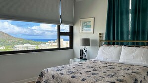 Un dormitorio con cama, mesita de noche, lámpara, un cuadro enmarco y una ventana que ofrece vistas a edificios y una colina.