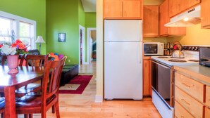 A compact kitchen with wooden cabinets, a white refrigerator, a microwave, and a stove. There is a dining area with a wooden table and chairs, and a red rug on the floor.