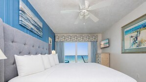 A bedroom with a large bed, a ceiling fan, a view of the ocean, and a painting on the wall.