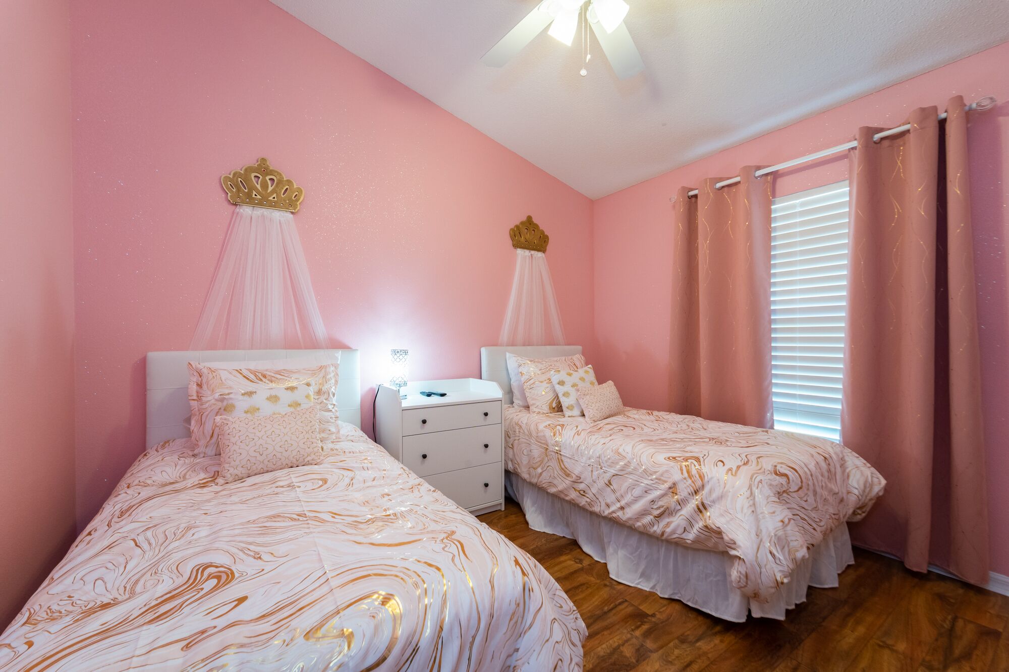 Une chambre à coucher avec deux lits, chacun doté d’un baldaquin et d’une décoration en forme de couronne, de tables de chevet blanches et de rideaux roses.