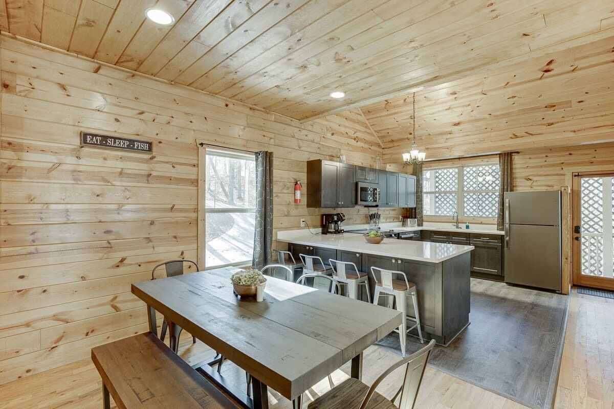 Interior de una cabaña de madera con comedor, cocina y grandes ventanales.