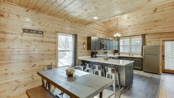 Interior de una cabaña de madera con comedor, cocina y grandes ventanales.
