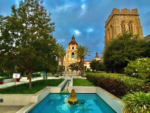 A pool with a statue in the center, surrounded by greenery and a building with a dome and tower in the background.