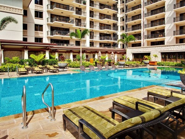 A hotel pool area with lounge chairs, a swimming pool, and a multi-story building with balconies.