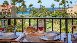 Una mesa de comedor con una preparación de comida, que incluye una ensalada, pan y copas de vino, con vista a un paisaje tropical con palmeras y vistas al océano.