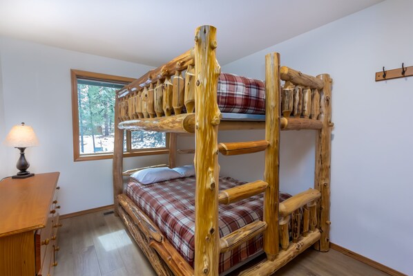 Una cama literera con colcha escocesa, un escritorio de madera con lámpara y una ventana con vista a árboles.
