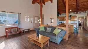 Ein geräumiges Wohnzimmer mit Holzboden, einem großen Fenster mit Blick auf eine verschneite Landschaft, einer Couch mit Kissen, einem Holztisch und einer Küchenzeile im Hintergrund.
