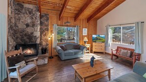Ein gemütliches Blockhaus-Interieur mit Kamin aus Stein, Holzbalken, einer Couch, einem Holztisch und Blick ins Freie durch große Fenster.