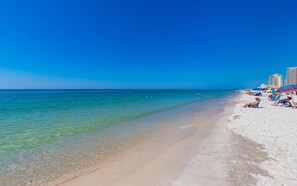Une plage avec une eau turquoise et limpide, du sable blanc et quelques personnes qui prennent un bain de soleil. Des parasols et des chaises de plage sont disposés le long du rivage.
