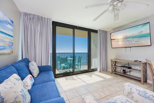 Un salon avec un canapé bleu, un téléviseur et un balcon donnant sur la mer.
