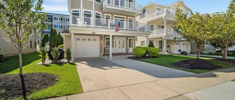 A multi-story residential building with a front garage, a balcony with a flag, and a well-maintained lawn.