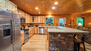 A modern kitchen with wooden cabinets, a stainless steel refrigerator, a stone island, and a view of the outdoors. - Budget-Friendly | Theater Room, Hot Tub, Gameroom (Gatlinburg)