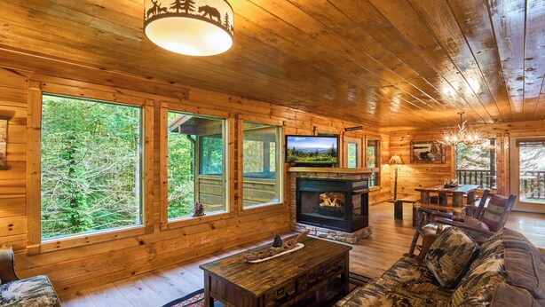 A log cabin interior with a fireplace, wooden furniture, and large windows overlooking a forest. - Budget-Friendly | Theater Room, Hot Tub, Gameroom (Gatlinburg)
