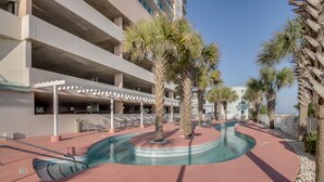 Zona de piscina con sillones de descanso y palmeras, rodeada por un edificio con balcones.