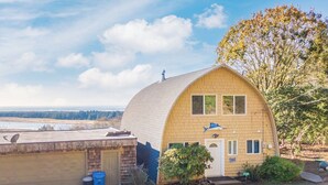 Une maison de plain-pied, dotée d’un toit incurvé, d’un garage, d’un petit porche à l’avant et offrant une vue sur une étendue d’eau en arrière-plan.