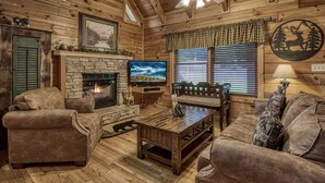 A cozy log cabin interior with a stone fireplace, leather sofas, a television, and a ceiling fan.