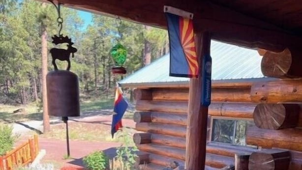 A wooden deck with a railing, logs stacked against the wall, and a flag hanging outside. - Hillside Rustic Retreat (Greer)