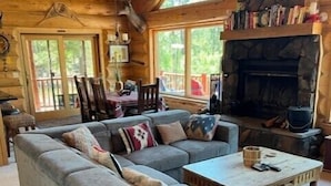 A log cabin interior with a large gray sectional sofa, a stone fireplace, and a dining area with wooden chairs and a table. - Hillside Rustic Retreat (Greer)
