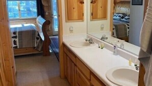 A wooden cabin bathroom with two sinks, a large mirror, and a window leading to a bedroom. - Hillside Rustic Retreat (Greer)