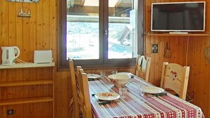 A wooden interior with a dining table set for four, a television mounted on the wall, and a window with a view of a snowy landscape. - Studio/Cabin/Mezz 5 pers with shuttle, 600m from center - Le Grand-Bornand (Le Grand-Bornand)