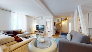 Una sala de estar moderna con un sofá, un sillón y una mesa de centro de vidrio. Un televisor está montado en la pared, y una escalera conduce a un nivel superior.