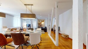 Un comedor amplio con una larga mesa dispuesta para una comida, rodeada de sillas modernas. En el fondo, un televisor está montado en la pared y una escalera conduce a un nivel superior.