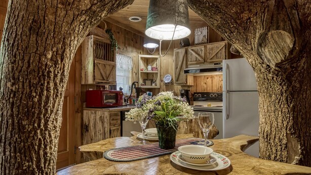 A rustic kitchen with wooden cabinets, a red microwave, and a dining table set with plates and a vase of flowers. - ER69 - Rachel's Tree House - Great location. Close to all the action! (Pigeon Forge)